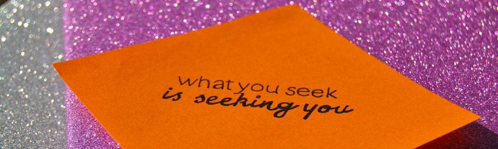 A sticky note with the inscription ‘What you seek is seeking you’ on a pink glitter background.