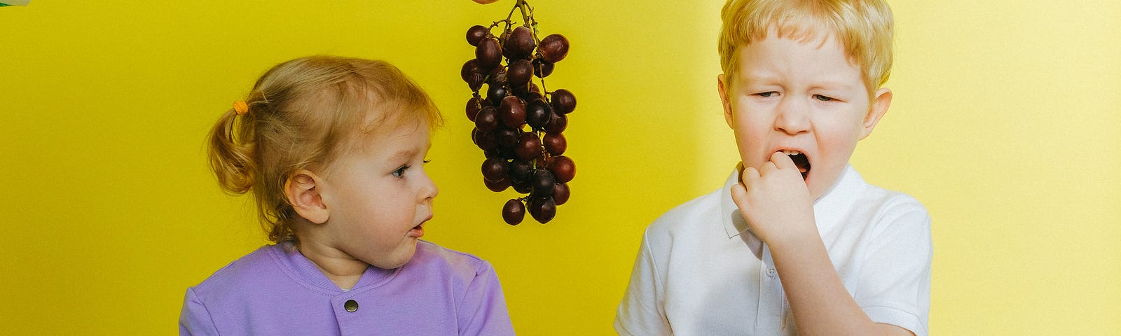 Two children. A hand from an off-camera person is offering grapes to the child on the right, but not the child on the right.