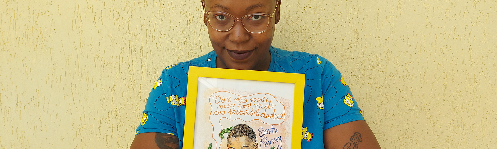 Na foto, uma mulher negra, careca, com uma camiseta azul, segura uma quadro escrito: você não pode ter medo das possibilidades