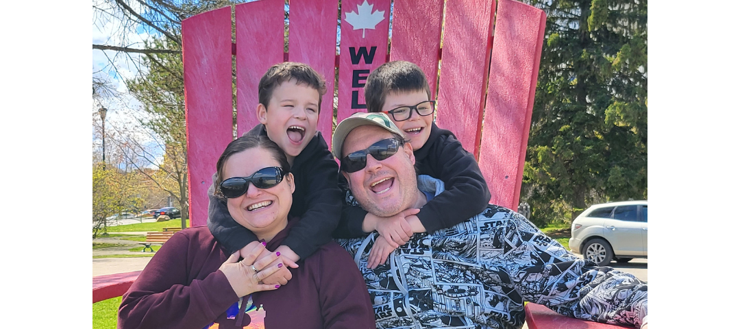 Photograph of the Author on the left; her partner on the right; and kids on a large red Adirondak style wooden chair outside on a sunny day