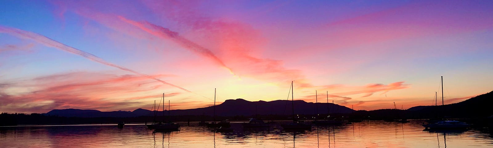 A sunset sinks over a body of water. The sky is blue and shades of peach and pink which is also reflected on the ocean below.