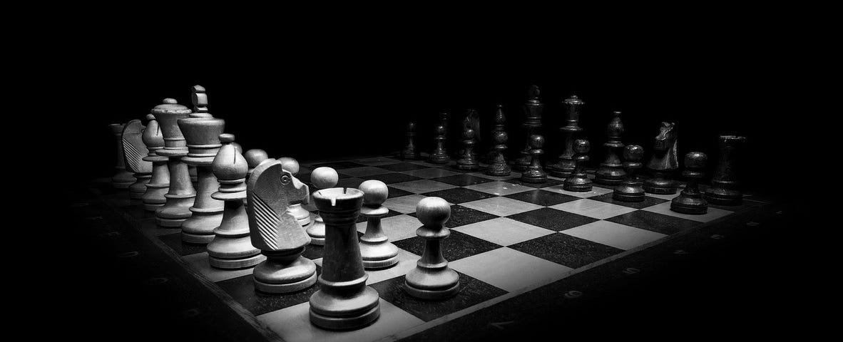 Image: A black and white chess set set out in deep and dramatic shadows. No one has yet taken a move.