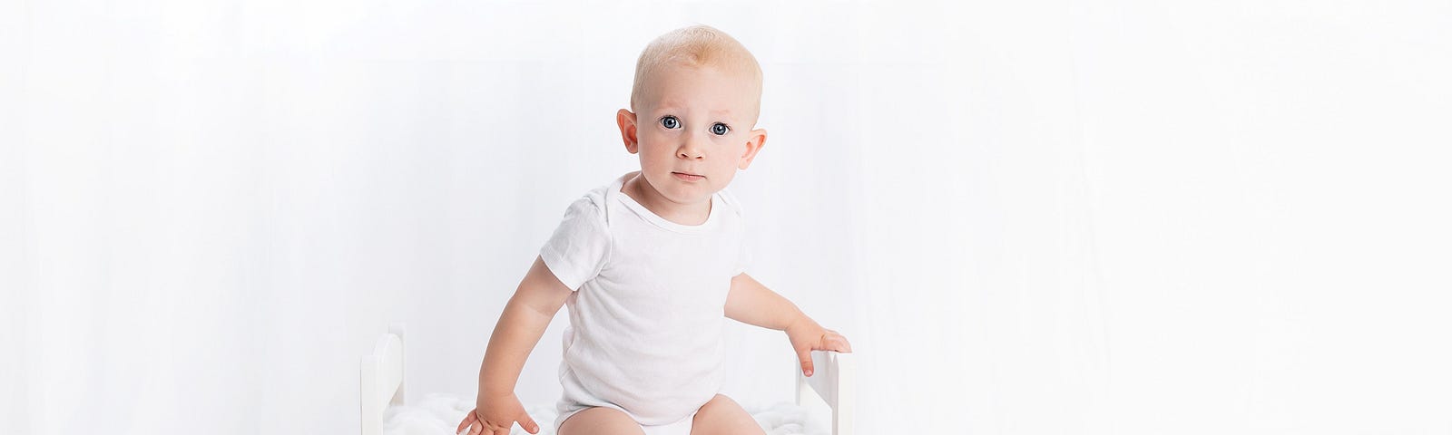 Toddler wearing a white tshirt and white underpants, sitting on a white chair in a white room