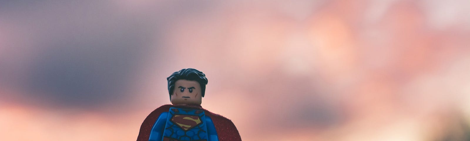 Closeup photo of a tiny Lego superman micro action figure, posed on top of what looks like a stump or wooden post, with a blurred background of sunset-or sunrise-tinted clouds behind