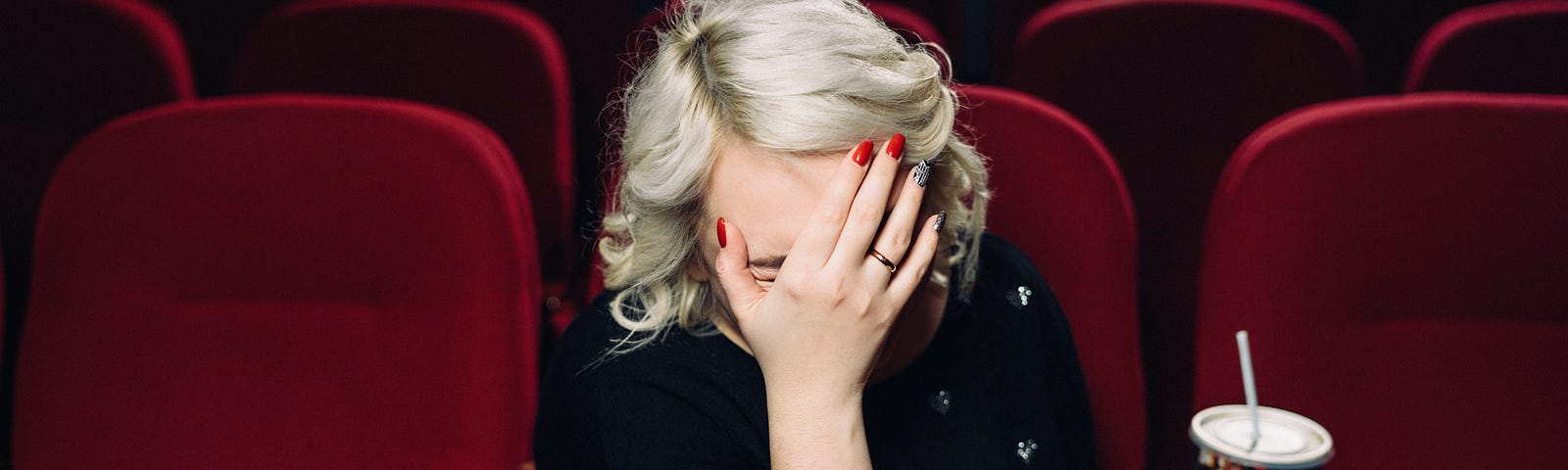 A blond woman in a movie theater palms her face with her hand. A tub of popcorn is in the foreground.