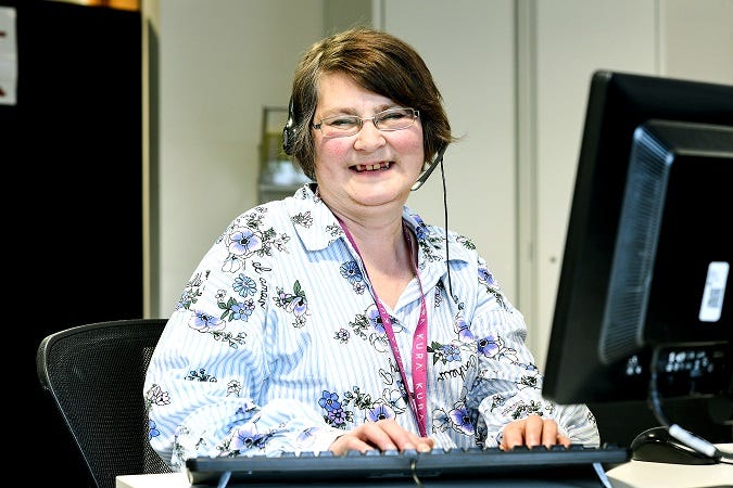 A woman smiles at the camera. She’s sat a desk in front of a computer with her hands in typing position on a keyboard. She wears a headset with microphone.