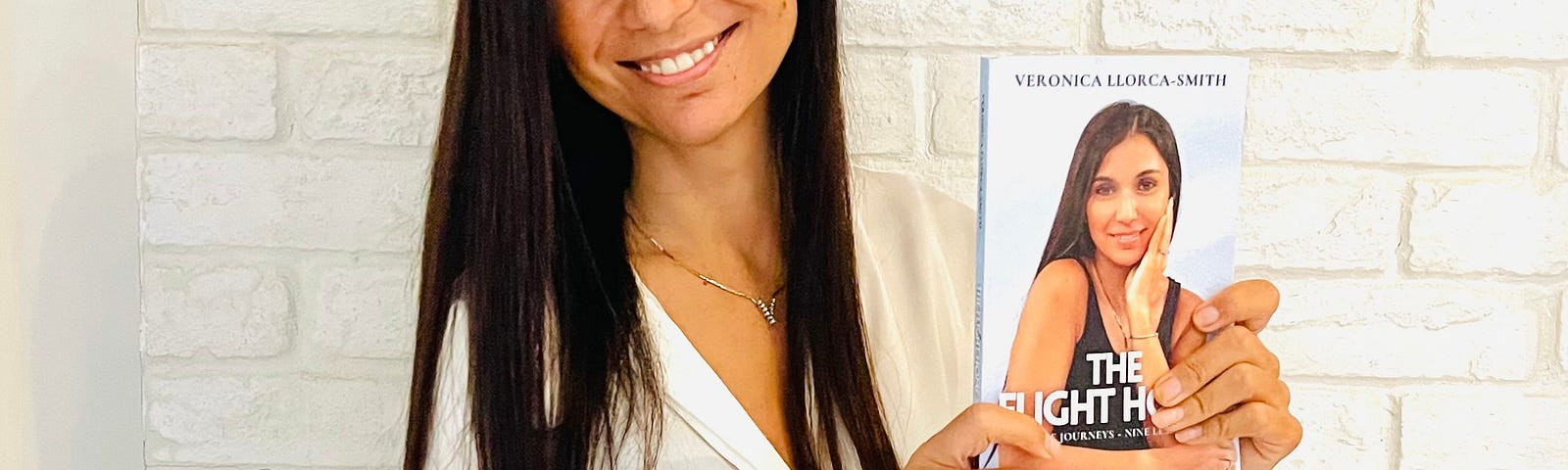 Woman with long black hair and a white blouse holding her book, the Flight Home. She’s smiling at the camera