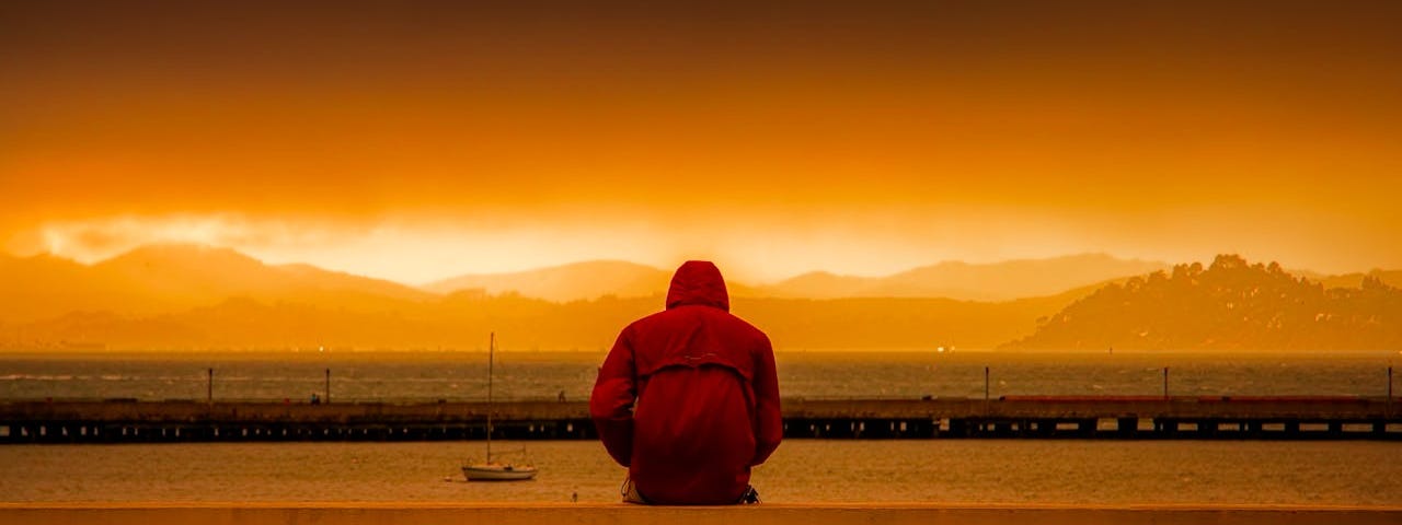 A person in a red jacket looks out at an orange sunset over water.