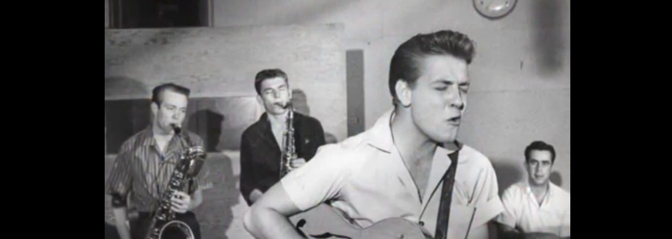 Eddie Cochran playing guitar and singing with two Saxophonists and a drummer behind him.