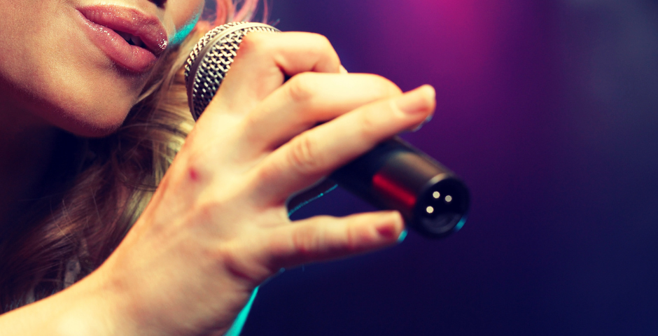 Singer sings into microphone held by their hand. Only their mouth is visible. Dark background with purple hue.