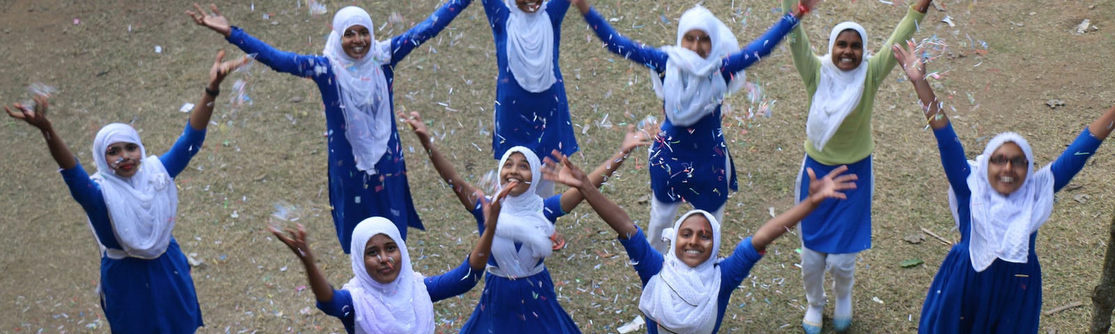 Bangladeshi girls in a blue school uniform and white headscarves throwing up their hands in a celebratory manner with confetti