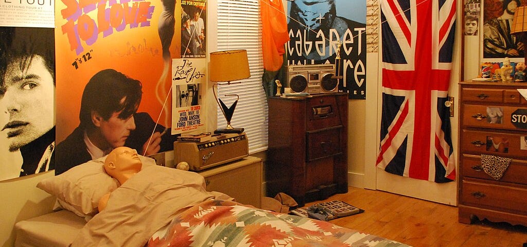 A bedroom with walls covered in posters, radio on a dresser, and a mannequin in a bed.