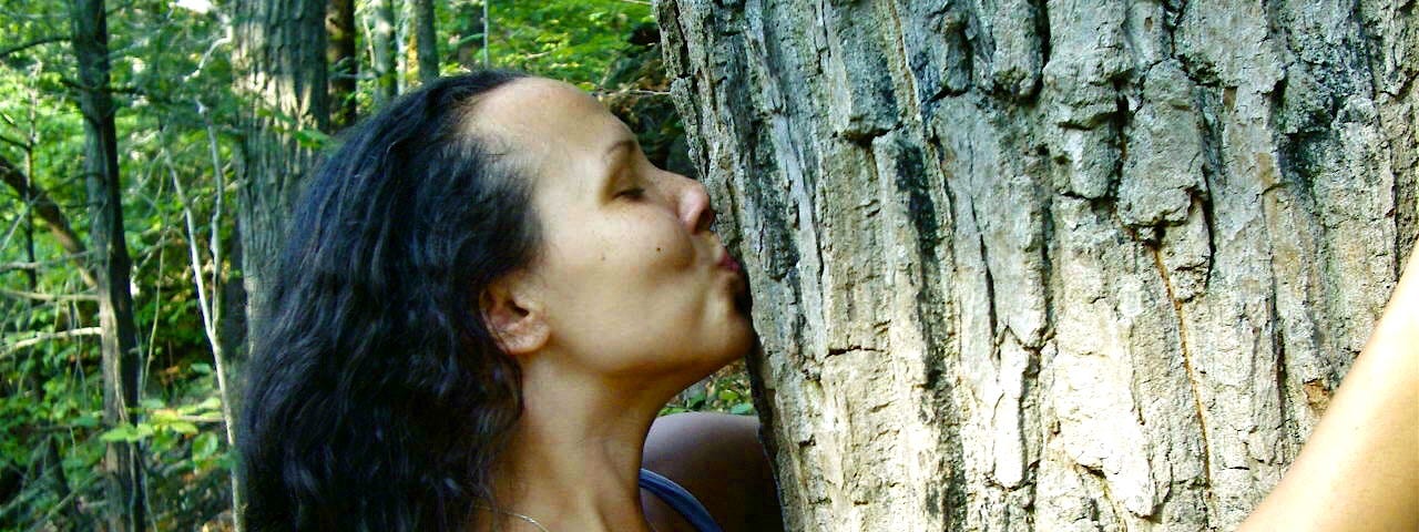 Profile of a woman with her arms wrapped around a tree while kissing the tree. Her eyes are closed.