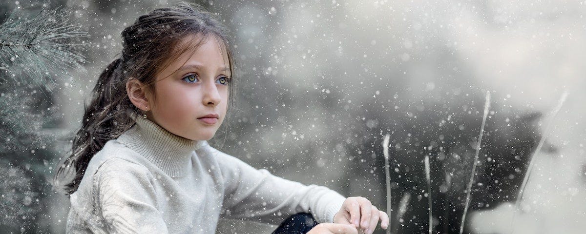 A pre-teen girl with dark hair and piercing green eyes sitting in the snow beside a fir tree. She is wearing black leggings and a white turtleneck sweater.