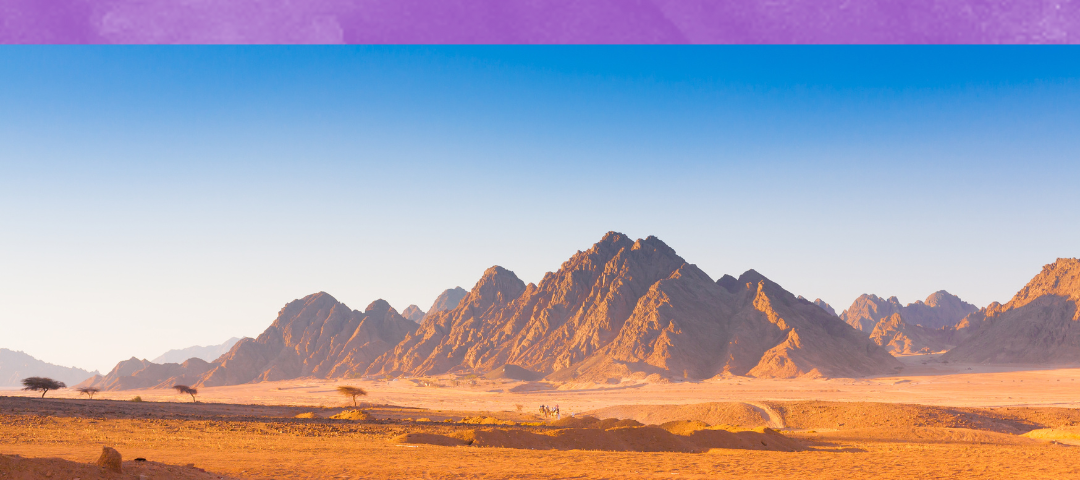 A panoramic view of a jagged mountain range from the point of view of a mile away in an orange-dirt desert.