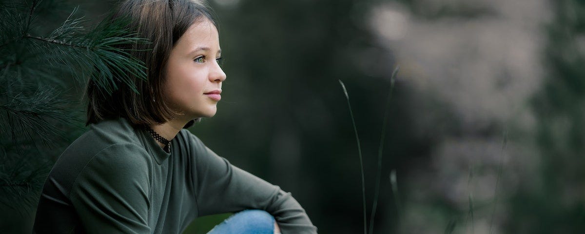 A young girl, perhaps nine years old, with intense, watchful eyes, sitting on grass beside a conifer. She has holes in the knees of her jeans.