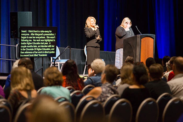 A scene from a conference presentation showing the audience sitting, a speaker presenting, and two accessibility supports: a sign language interpreter and a real-time captioning screen.