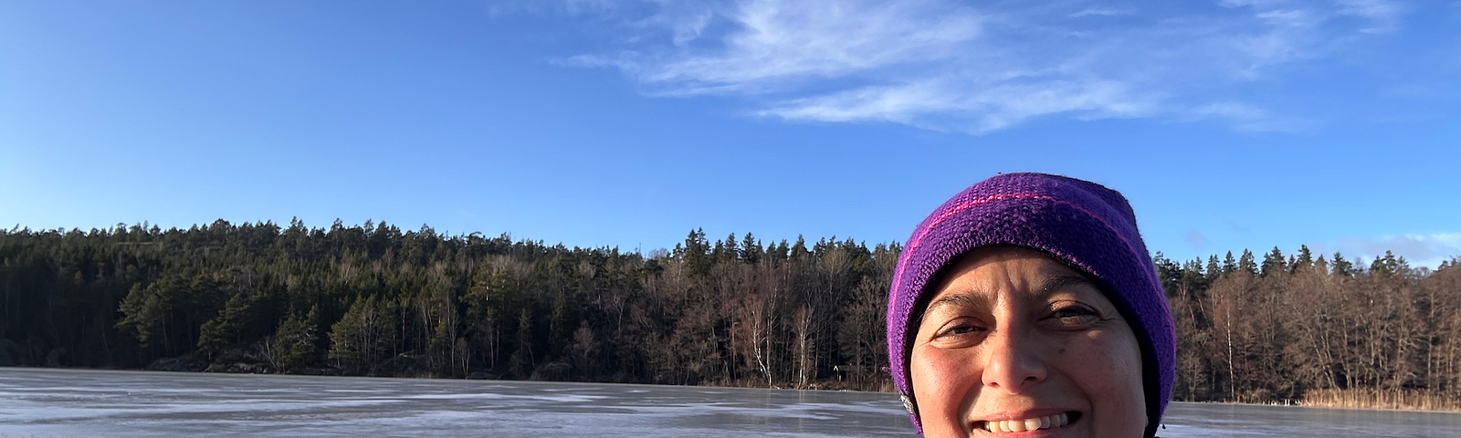 Zayera is wearing purple hat and winter coat. Blue skies and forest in the background. Hiking on a frozen ice lake in Stockholm region in Sweden. Zayera is smiling and looking into the camera.