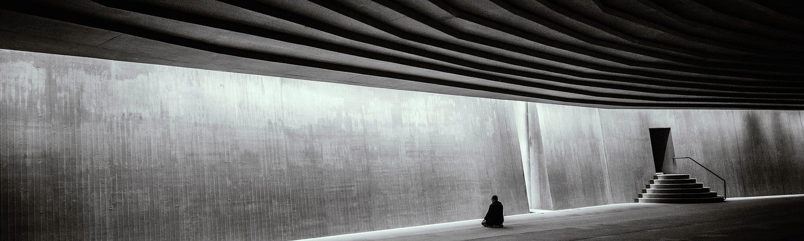 Black and white. A figure in the distance of a concrete empty hall sitting in meditation.