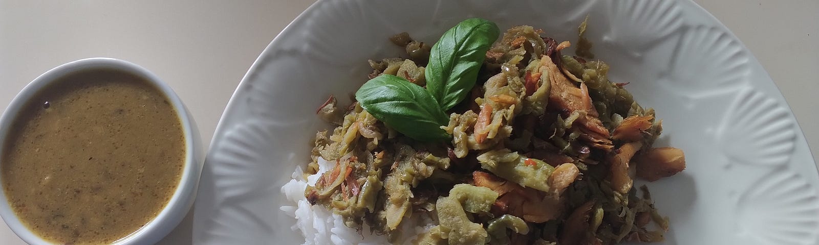 A white dish plated with white rice, green cooked karela aka bitter melon and decorated with cherry tomatoes, halved and arranged to replicate a red flower. On it’s left side a tiny bowl of green dahl soup a delicious addition to the meal.