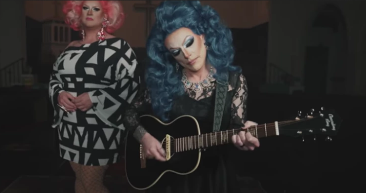 Two people in drag. The one in the back is wearing a black and white patterned dress and a pink wig. The one in the front is wearing a black lacy dress and a blue wig and playing a guitar.