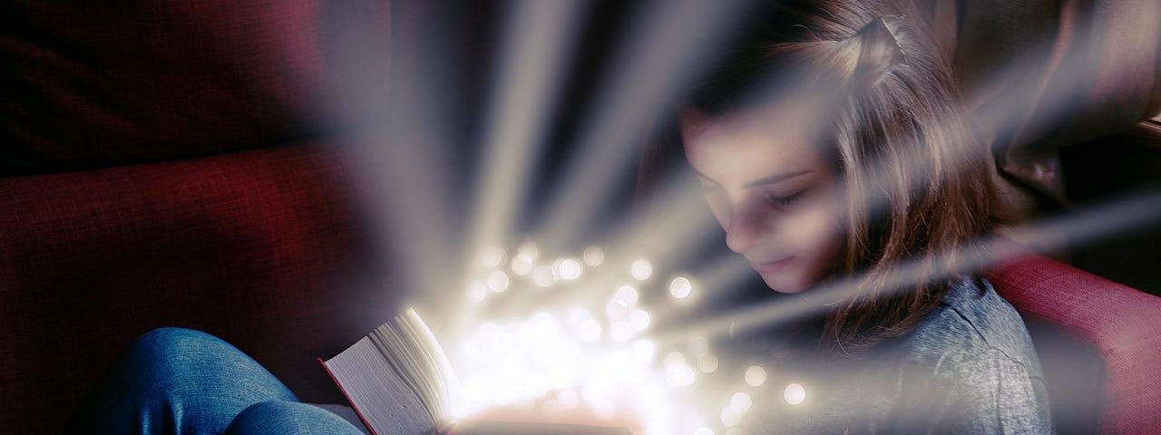 A young girl sitting, reading a book. The book is open on her lap and the magic is surrounding her in rays of light and glitter light.