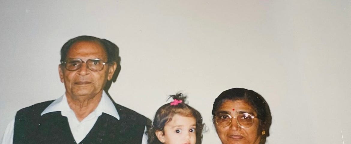 A baby, middle, wears a pink outfit and sits in the arms of her grandmother, a dark-skinned woman with a black bun and colorful saree. A man, the baby’s grandfather, stands to the left, wearing a white collared shirt and black vest.