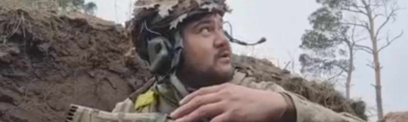 Ukrainian soldier in full military gear, looks up toward sky, from a ditch or place of hiding.