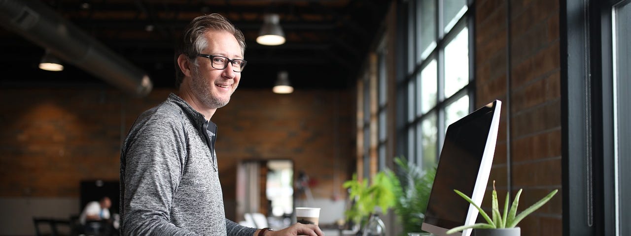 A middle aged man sits at a desk facing a window. There is a small plant, some books and a computer screen on his desk. He wears glasses and a long sleeved button down shirt. He is smiling.