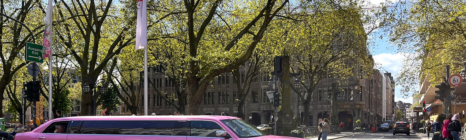 A bright pink limousine about to go across an intersection on an urban street with trees in the background on a sunny day.