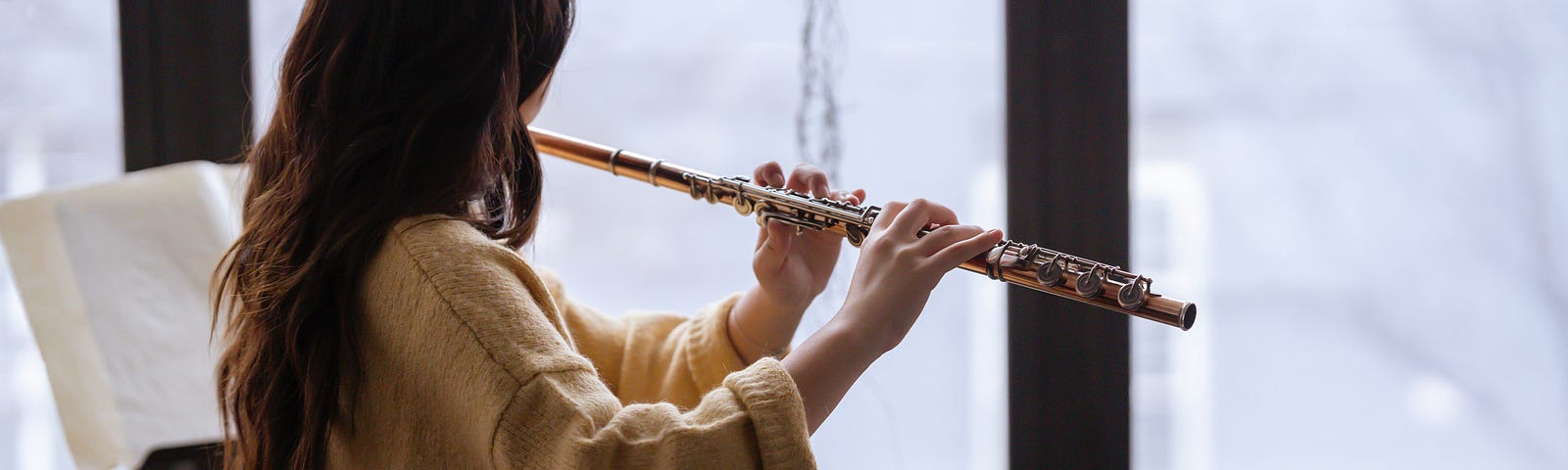 Photo of woman playing flute next to window in studio. She is facing the window, face away from the camera.