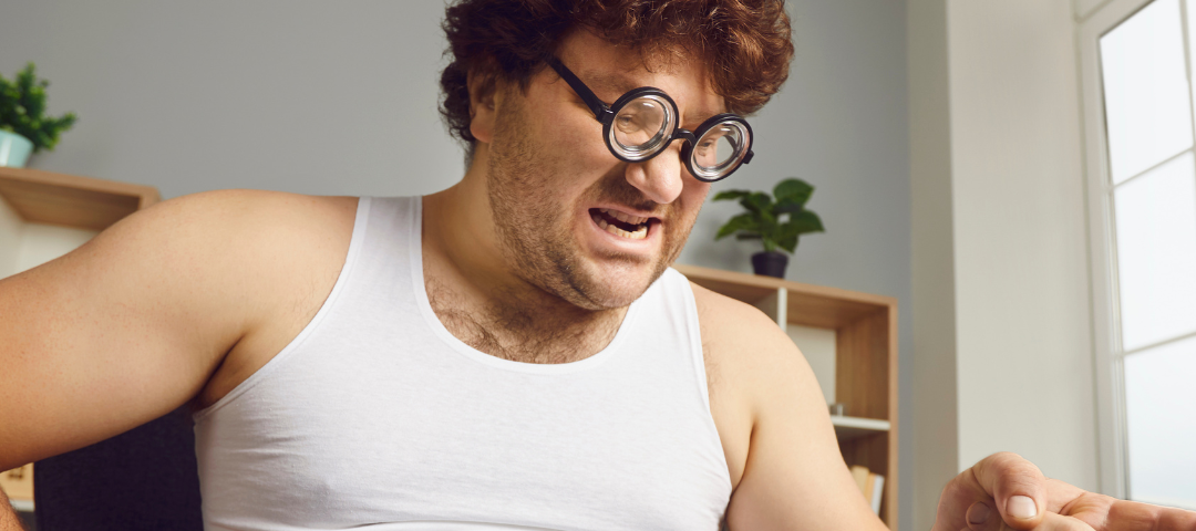 Man sitting in a white sleeveless undershirt and pj’s looks and points angrily at his computer.