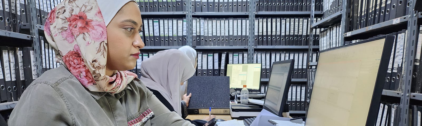 A woman types on a keyboard in front of a computer monitor inside an office with a few hundred matching black binders with white labels lined on shelving that nearly reaches to the ceiling.