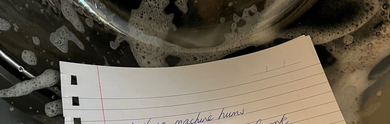 A washing machine in operation with a handwritten note with the words, ‘washing machine hums/ background  music while I work/ tedium repeats’, resting in the recessed part of the door.