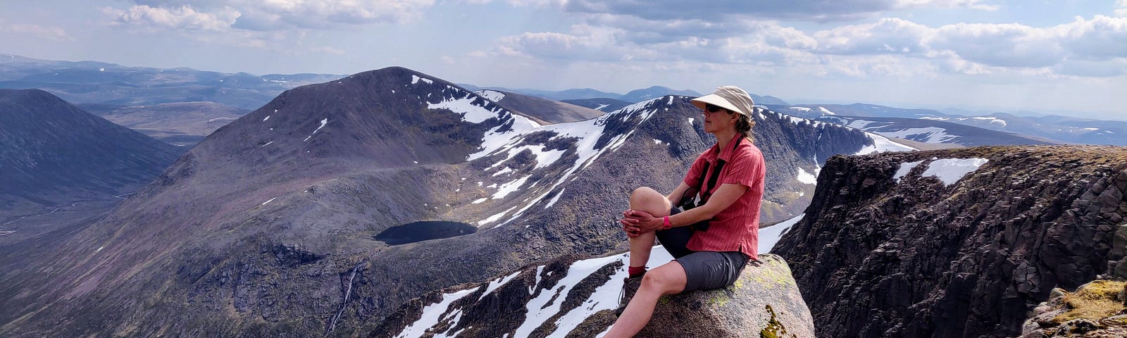Photo of author Merryn Glover wearing hat and sunglasses, relaxing in the Cairngorms with a stunning backdrop of the mountains’ snowy peaks behind her
