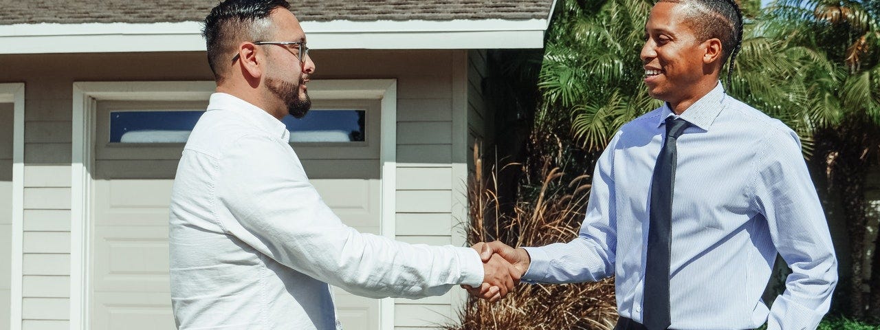Two men shaking hands in front of a house with a sold for-sale sign.