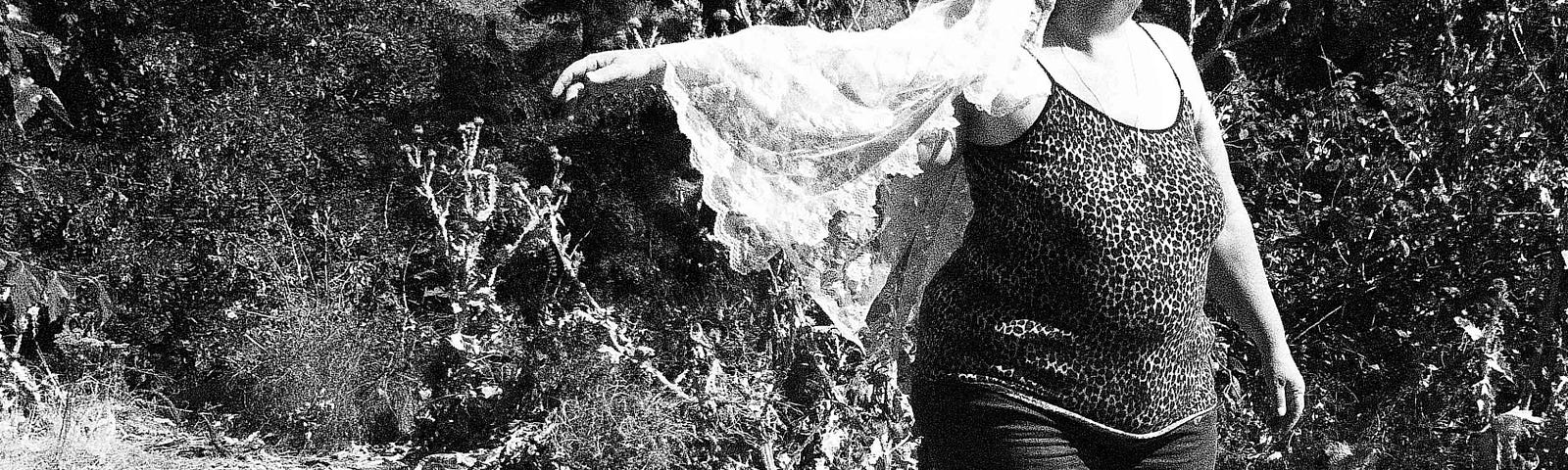 Black and white photo of a fat woman wearing cutoff shorts, leopard print spaghetti top, wedding veil posing out doors.