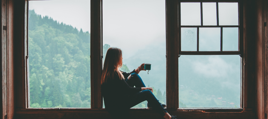 A woman holding a coffee cup gazing out the window into the mountains.