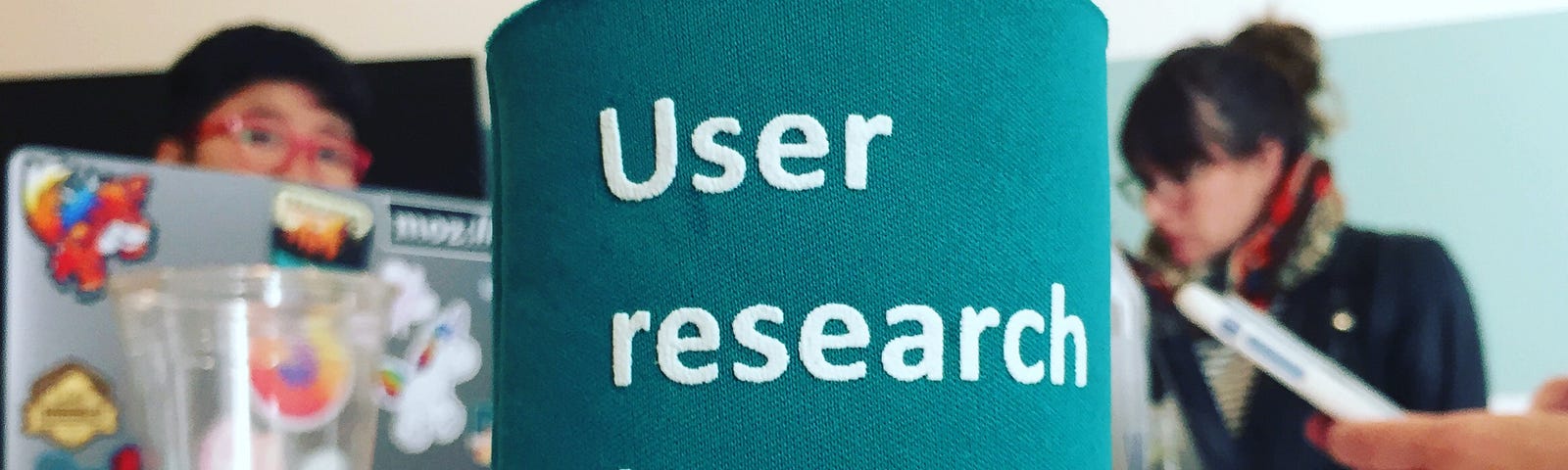 A soda can in a coozy that says “User research is a team sport,” sitting on a table with people & laptops in the background.