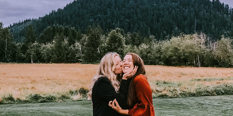 Cassie and Taylor stand in a grass field backed by trees. Cassie is kissing Taylor on the cheek.