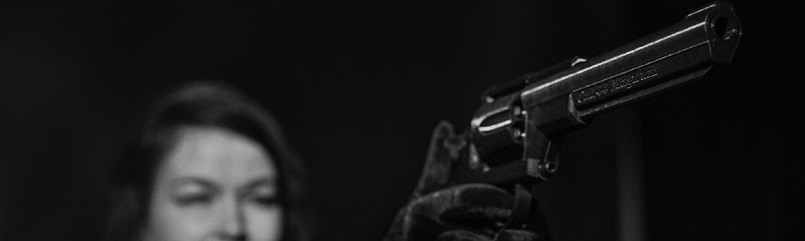 A black and white photo of a woman in an evening dress, wearing long velvet gloves. She holds a revolver in her hand and aims it at somewhere outside the picture. The camera focuses on the gun, while the woman’s face is out of focus.