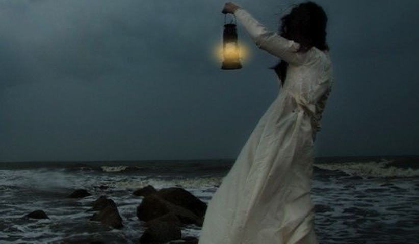 Dark-haired woman in a long white dress, standing on rocks and holding a lantern toward a dark sea.
