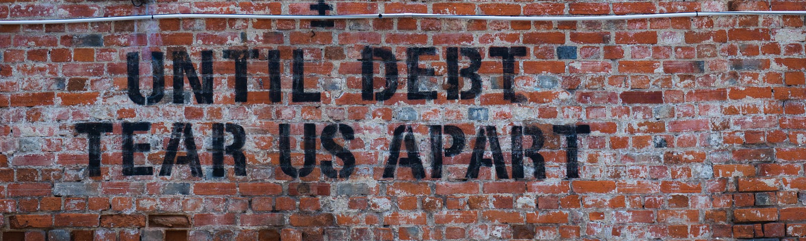 Red brick wall with a car parked in front of it. Text on the brick wall is black stencil grafitti that reads, “Until debt tear us apart” in all caps.