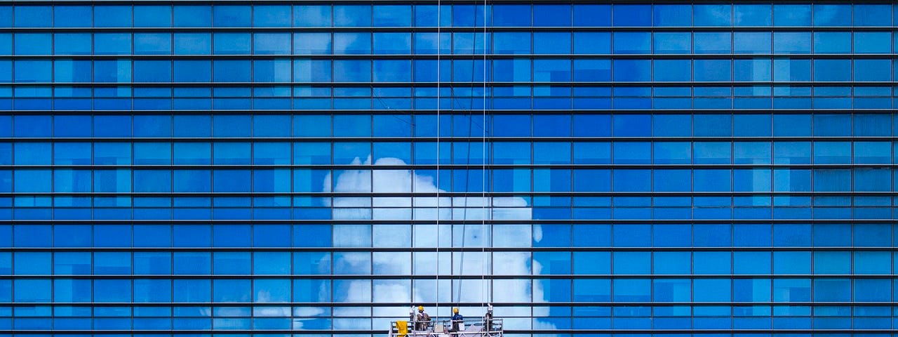 IMAGE: A huge glass skyscraper reflecting a blue sky with clouds, and workers hanging while they clean its façade