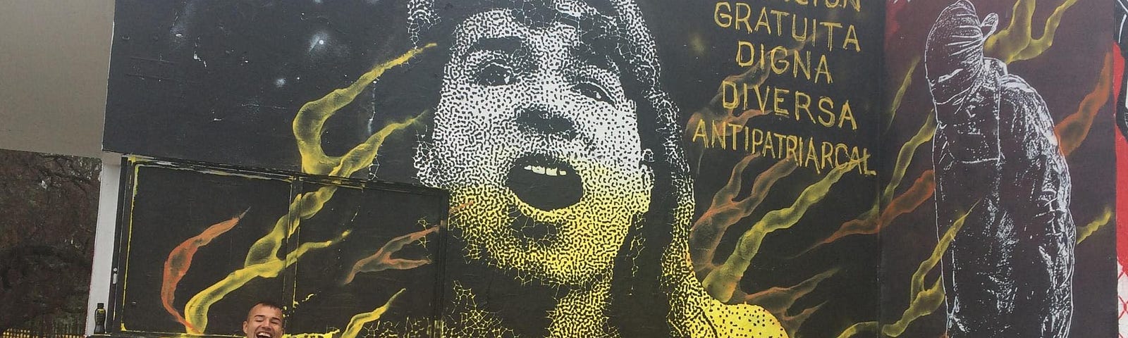 Una fotografía de Ammarantha frente a su propio mural en la Universidad Nacional de Colombia. Además de su rostro, el mural tiene una frase que dice “educación gratuita, diversa y antipatriarcal”.