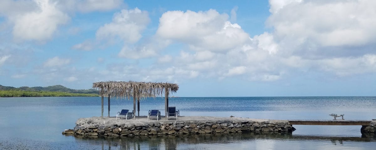 Empty lounge chairs beneath a large, thatched umbrella on the edge of a calm, blue bay