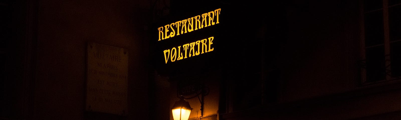 A yellow neon sign which reads “Restaurant Voltaire”, with a commemorative plaque to its left with information about Voltaire’s birth, and an old-fashioned street lamp underneath.