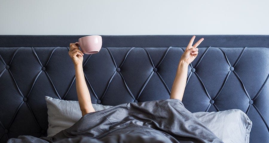 Woman in a large grey bed, head covered with a grey blanket- only her arms show- they are raised up- one with a large pink coffee cup and the other raising the peace sign