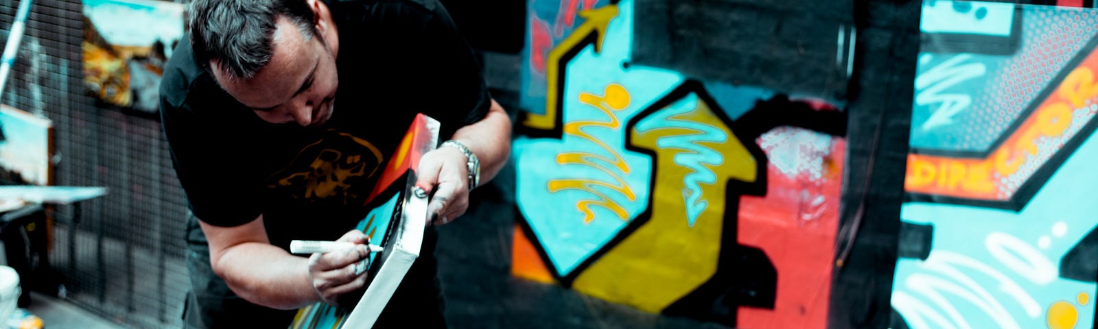 A street artist bending over to work on one of his peices. A collection of colourful artwork behind him.