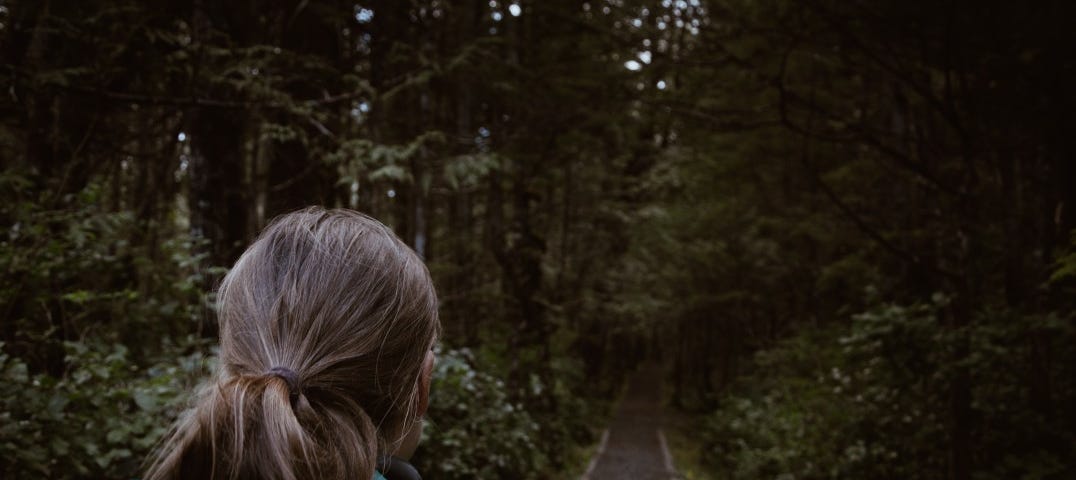 A woman stares down a dark wooded path with her back to the camera.